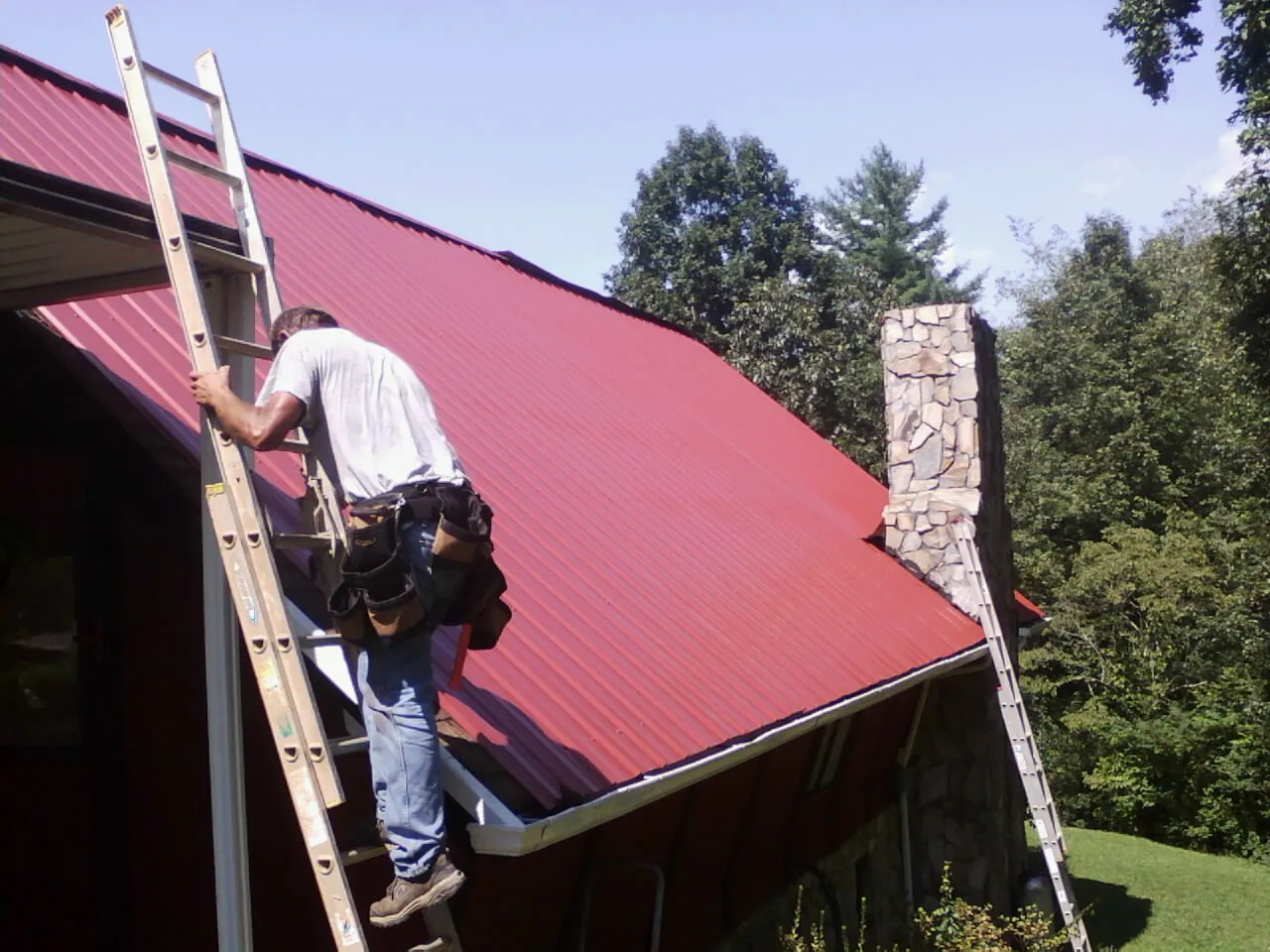 Roofer on a ladder beside a red metal roof and stone chimney — Soffit & Fascia Repair by Roofing Brothers in East Dennis